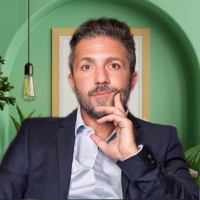 Homme barbu en blazer bleu marine posant pensivement devant un mur vert avec des plantes.